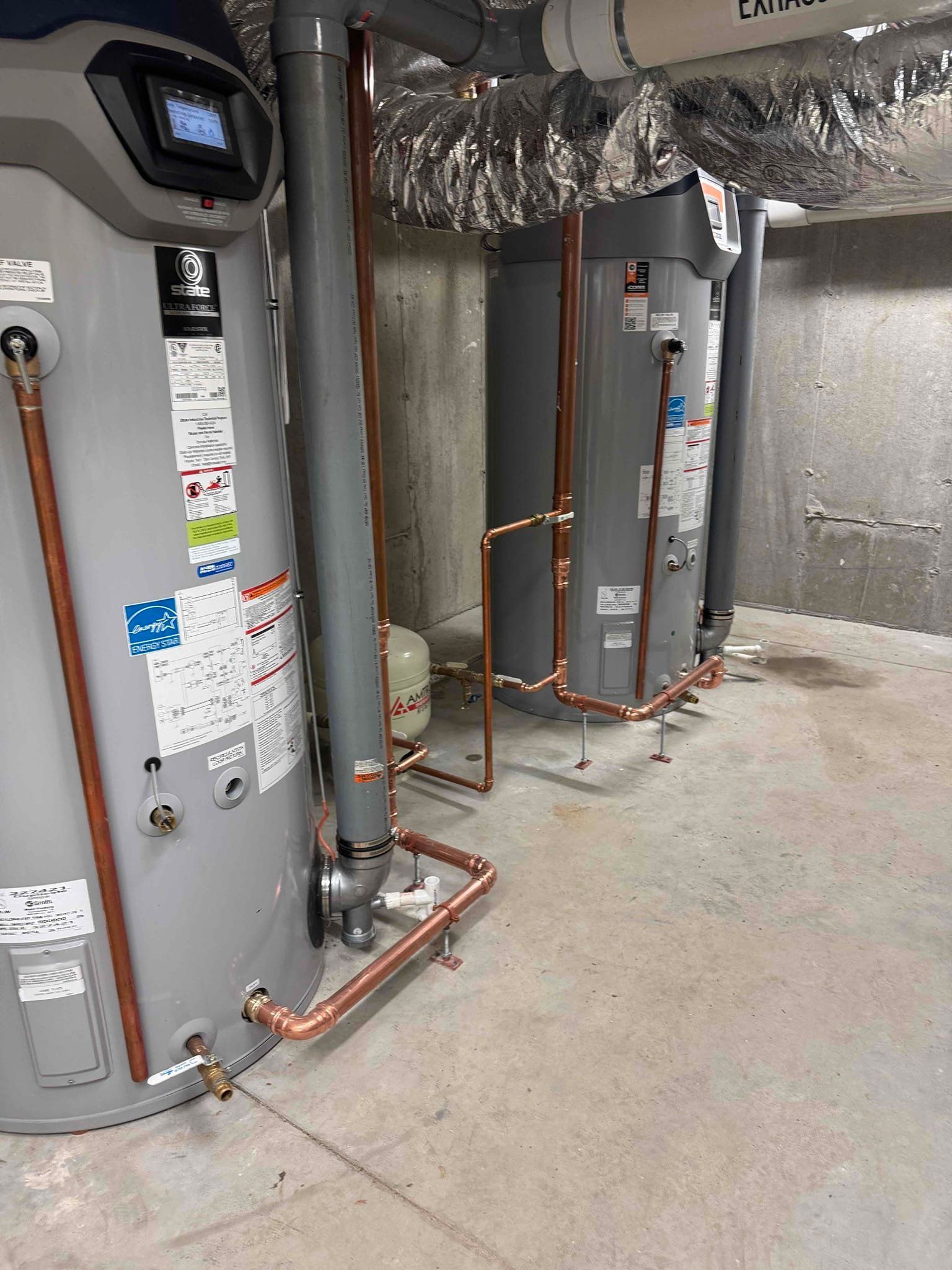 Basement utility room with large water heaters, pipes, and insulated ductwork against gray walls.