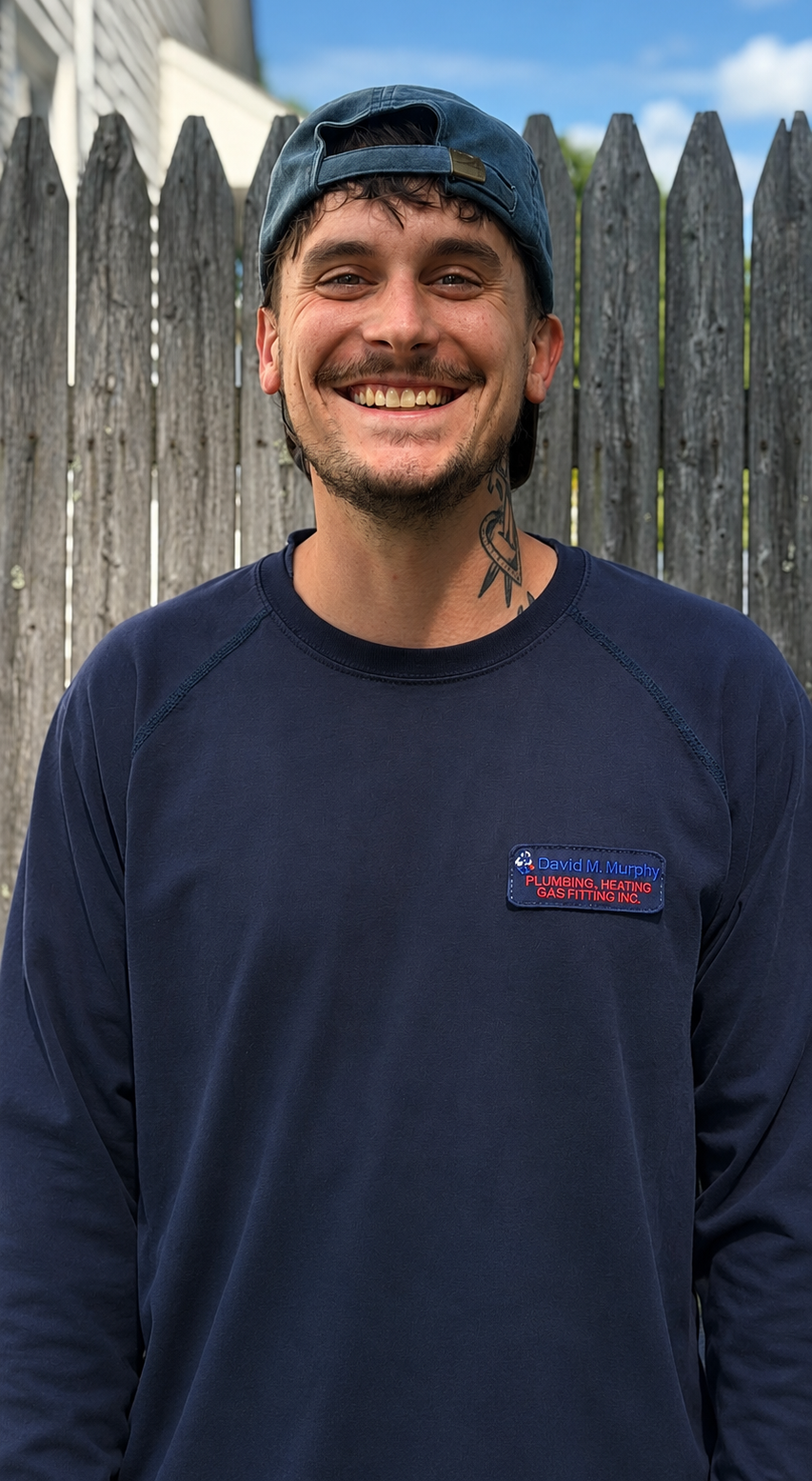 Smiling man in a navy shirt and cap standing in front of a wooden fence outdoors
