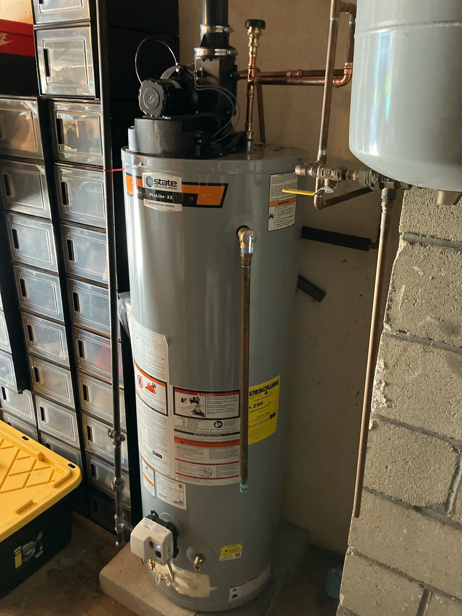 Tall gray water heater in a utility closet with pipes and a white tank above it