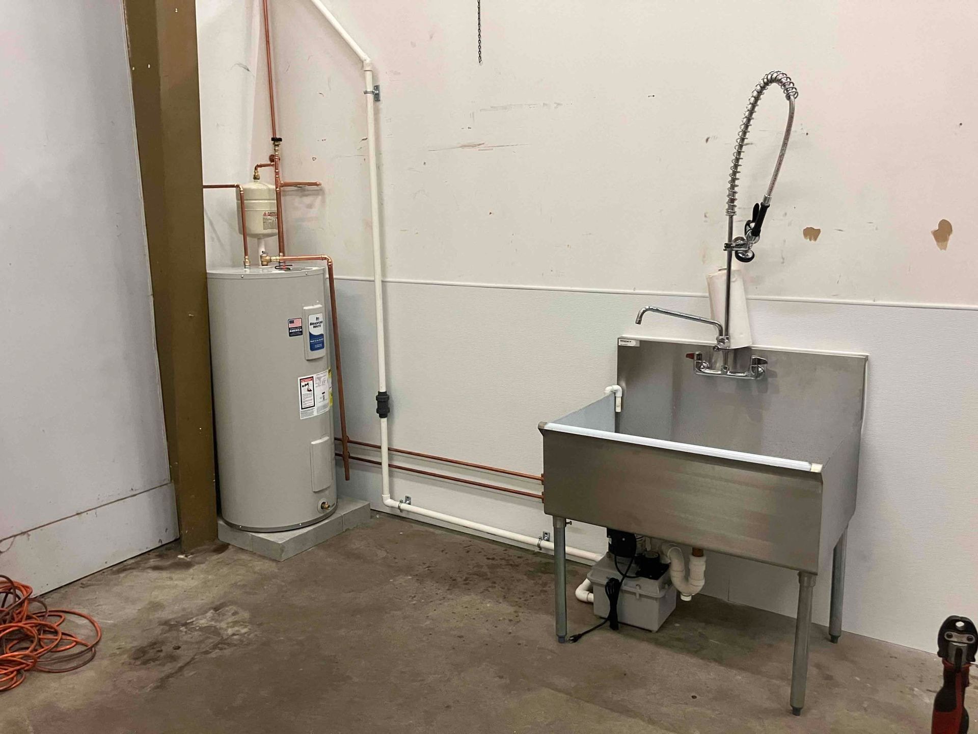 Utility room with a stainless-steel sink, water heater, and exposed pipes against a plain wall.