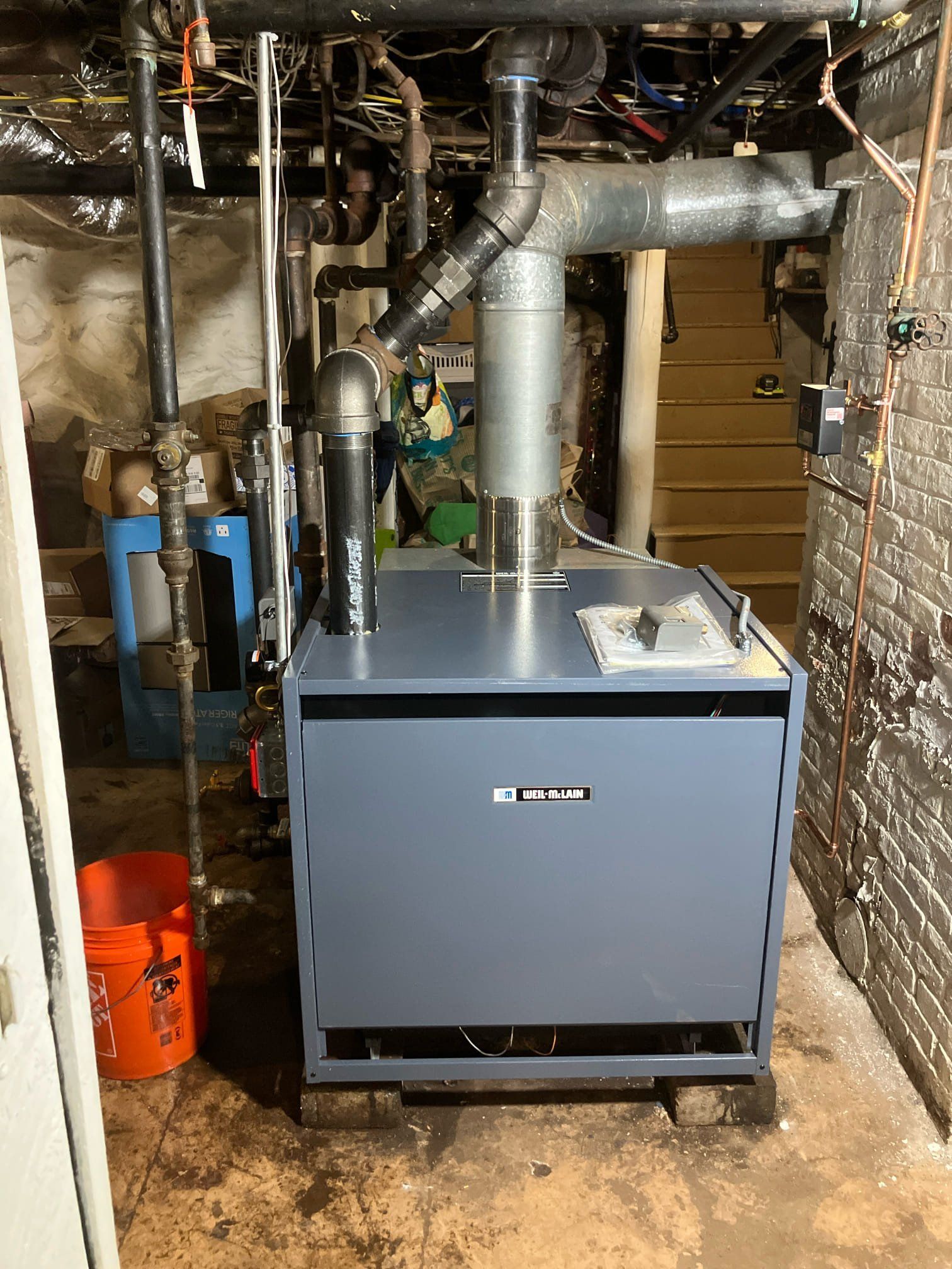 Basement furnace with silver ductwork and blue-gray unit against brick walls, surrounded by pipes and utility equipment