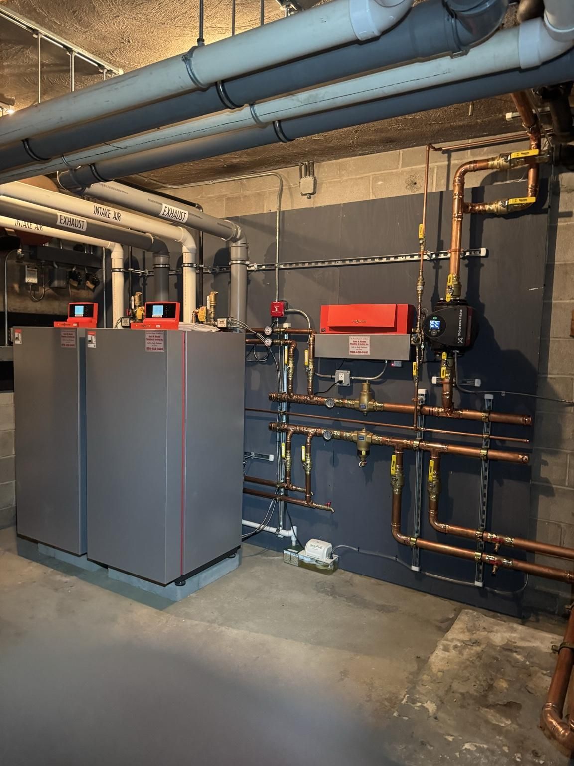Basement boiler room with gray and red heating units, exposed pipes, and industrial equipment.