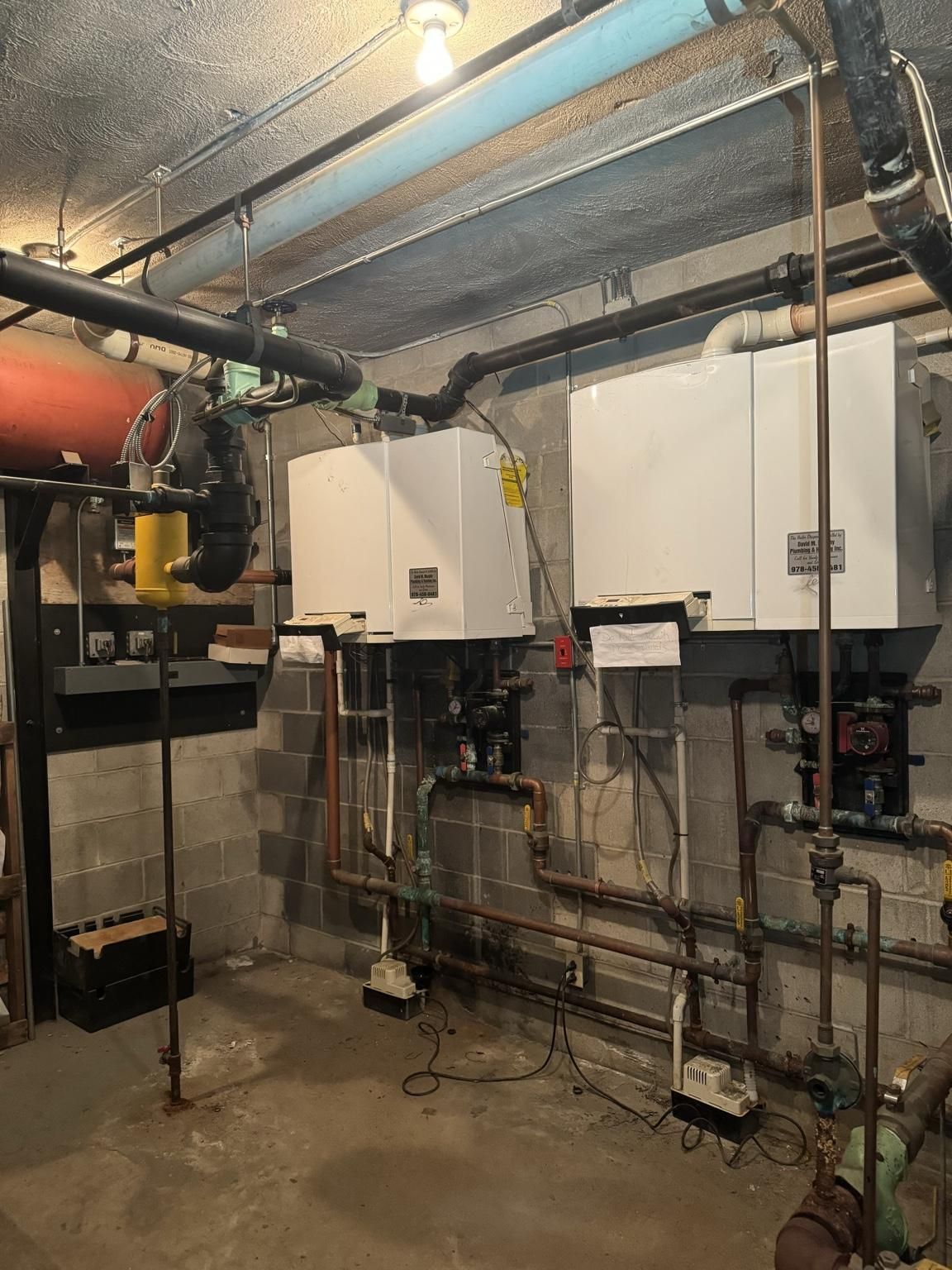 Basement utility room with wall-mounted boilers, exposed pipes, and electrical panels.