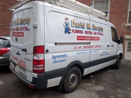 White plumbing service van with company logos parked on a street beside a brick building.