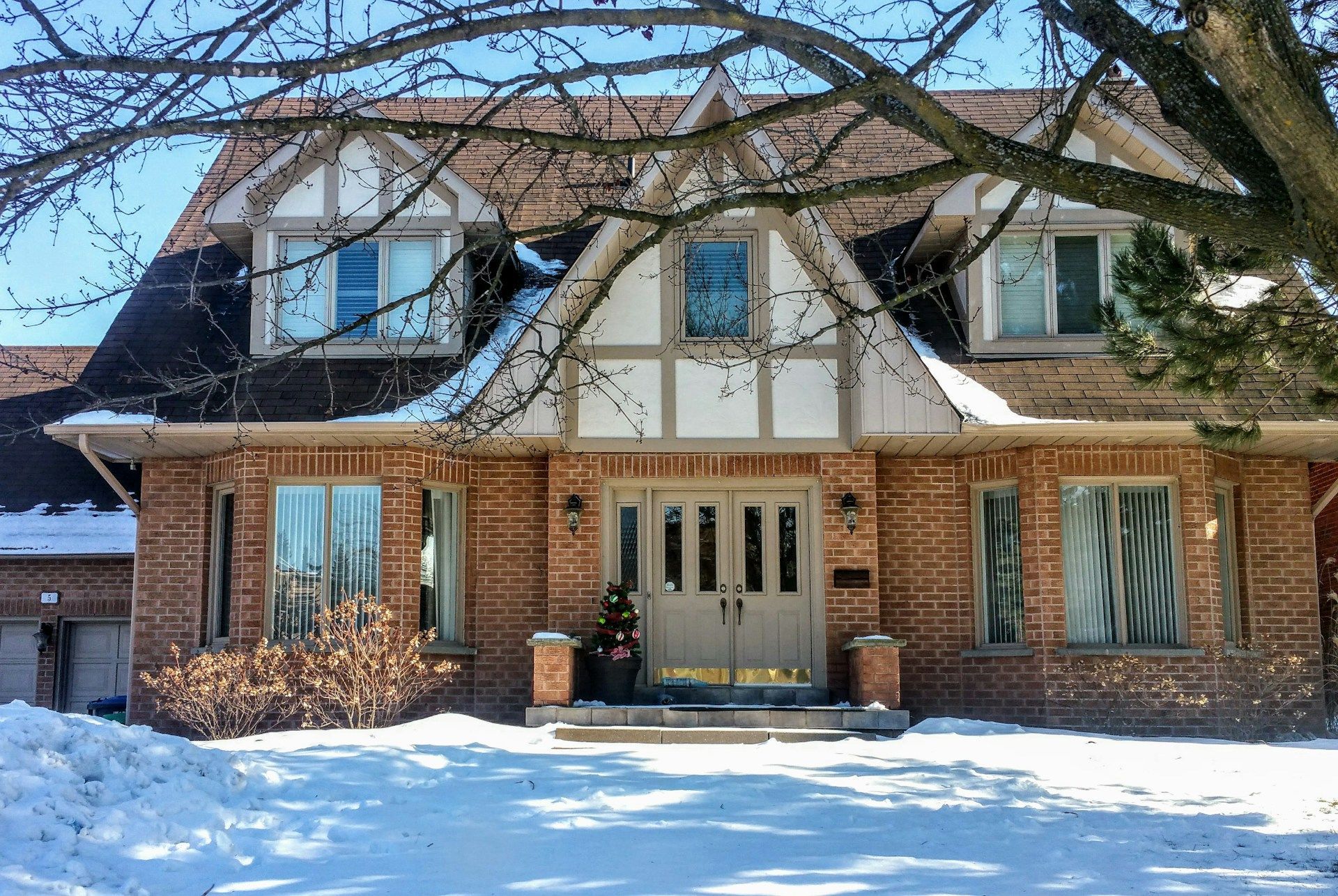 Snow-covered brick house with dormer windows and a central front door beneath bare tree branches