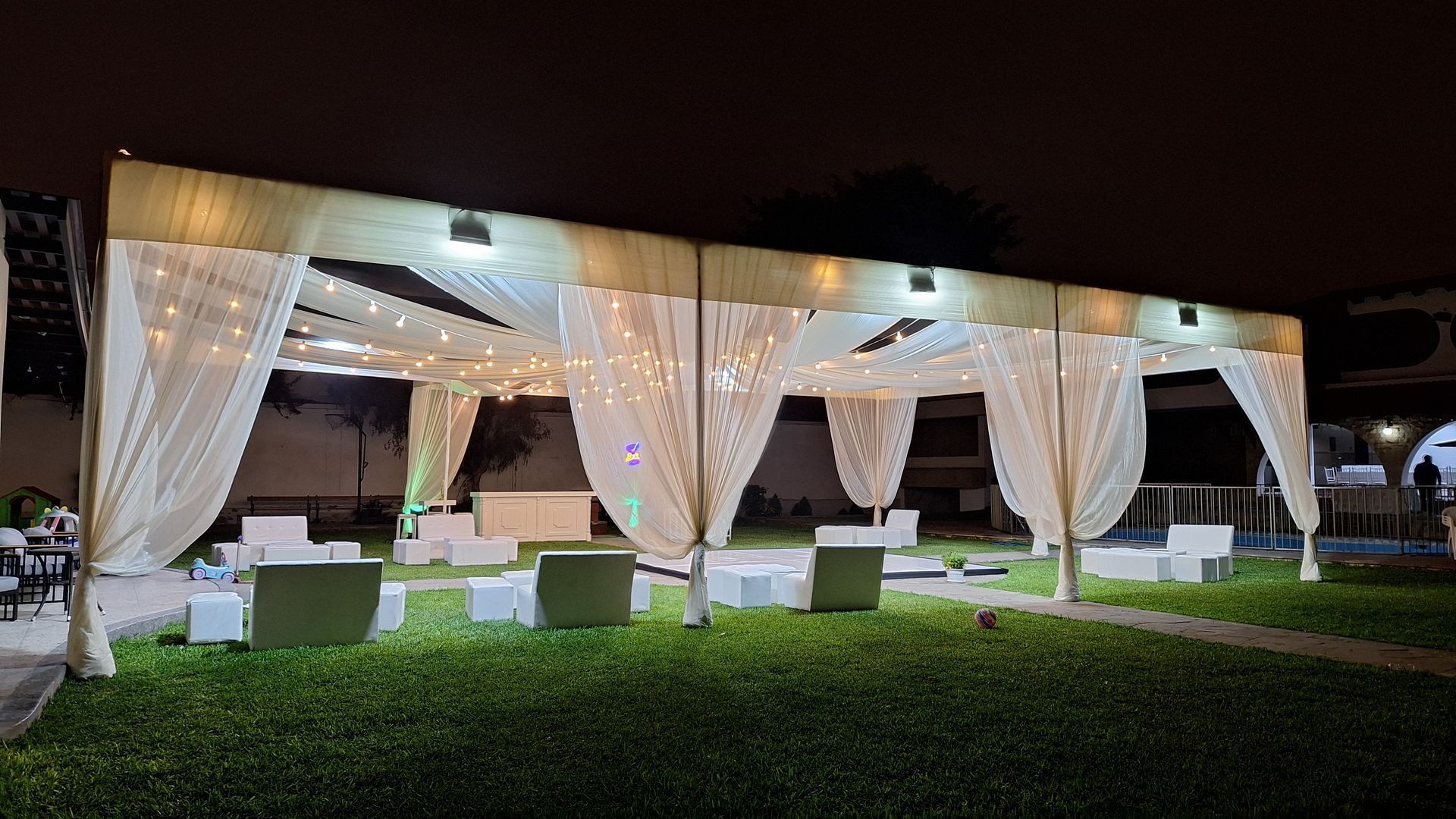 Salón de eventos al aire libre iluminado con cortinas blancas, sillas acolchadas y césped verde por la noche.