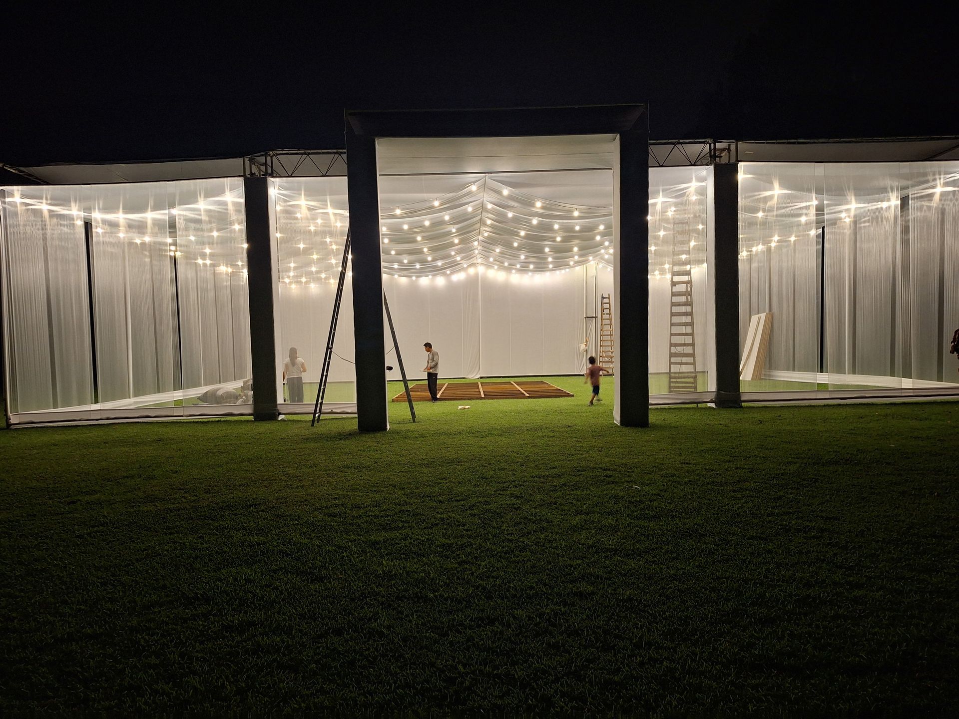 Guirnaldas de luces iluminan una carpa blanca para eventos por la noche, sobre un césped.