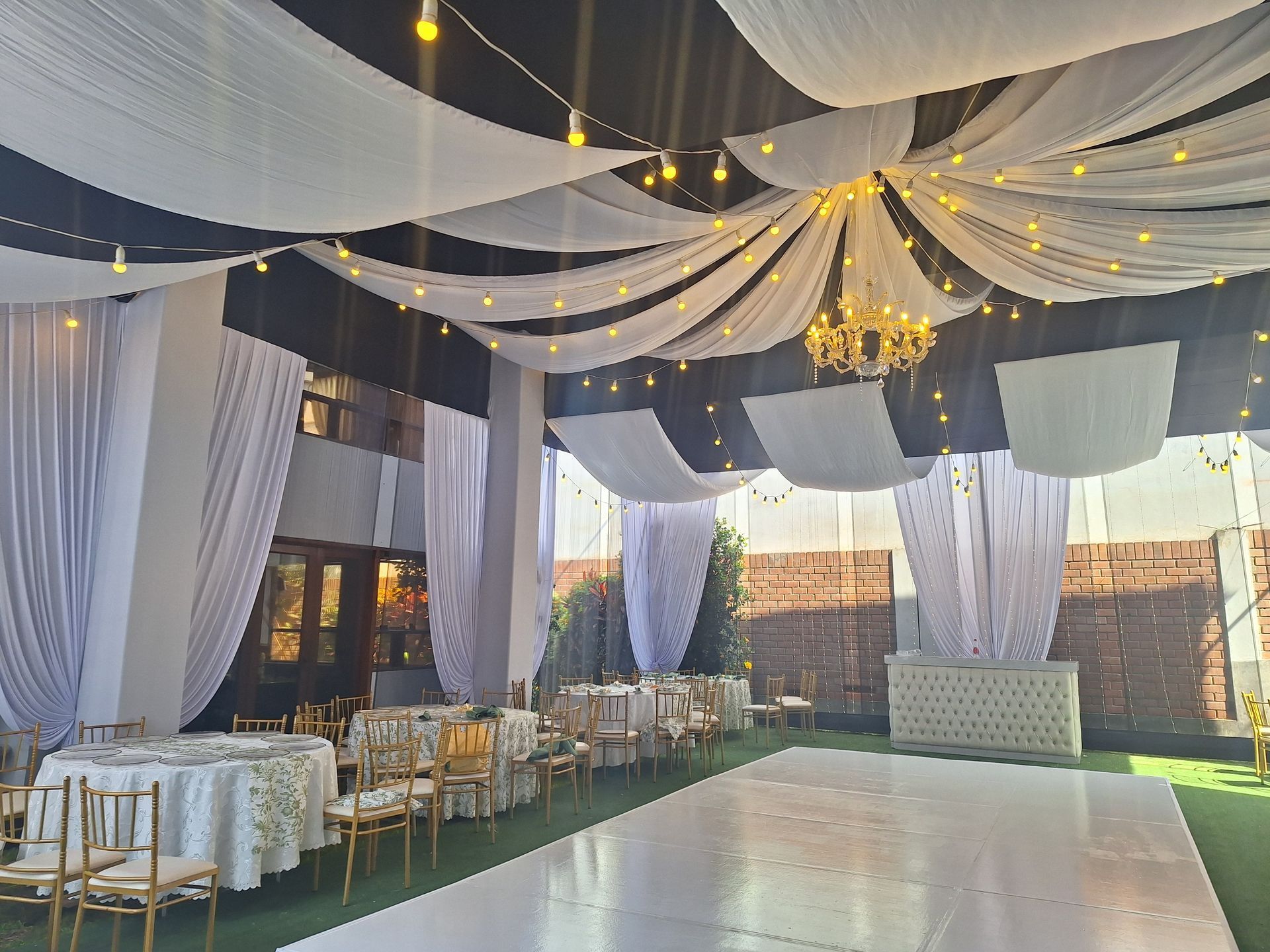 Elegante espacio para eventos al aire libre con cortinas blancas, luces centelleantes, candelabros y filas de mesas y sillas.