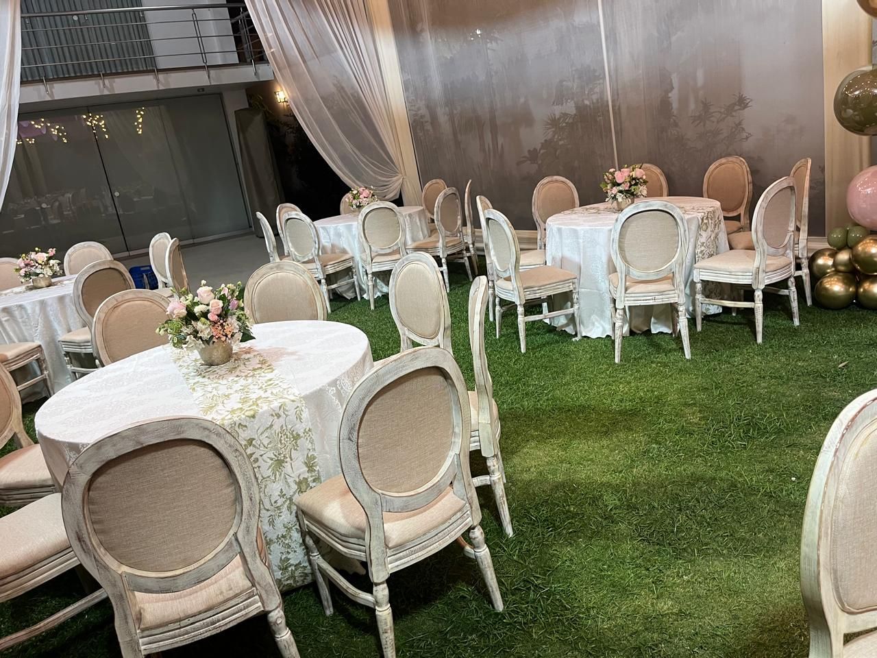 Montaje de evento al aire libre con mesas redondas y sillas blancas sobre un césped verde bajo cortinas transparentes.