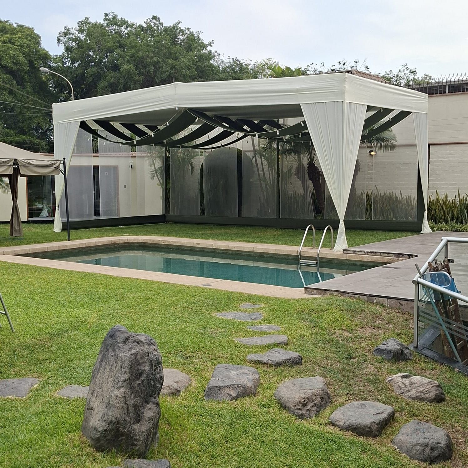 Piscina en el patio trasero, junto a un pabellón con toldo blanco, con césped verde y losas que permiten caminar sobre ella.