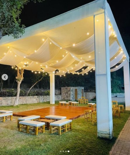 Patio exterior con toldo blanco, luces de guirnalda cálidas y asientos bajos de madera sobre el césped por la noche.