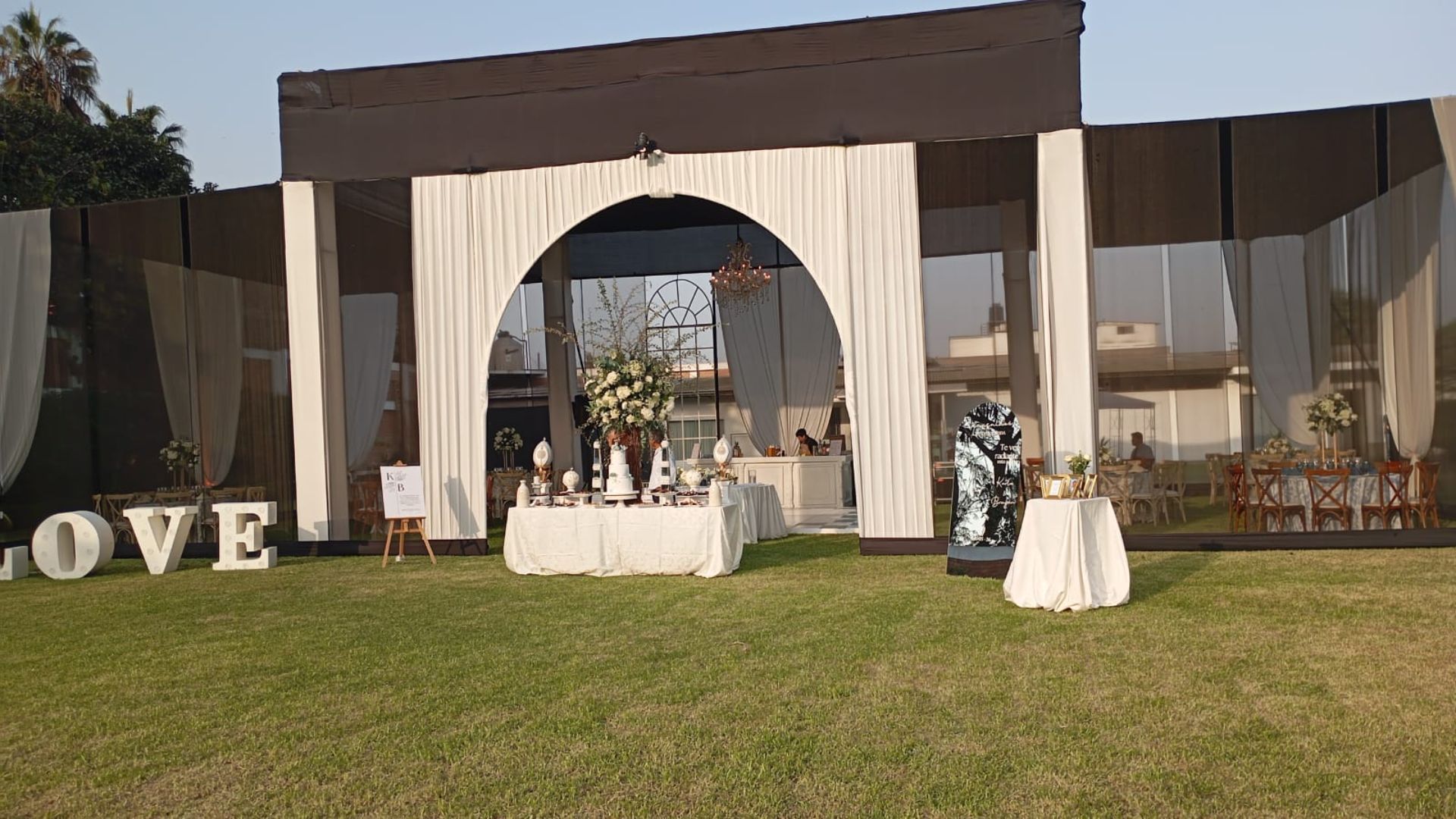Montaje de ceremonia de boda al aire libre con cortinas blancas, pasillo y decoración floral en un césped bajo un toldo.
