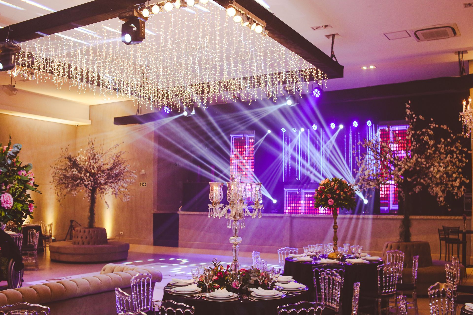Elegante salón de banquetes con luces de escenario moradas, candelabro de cristal y mesas decoradas para un evento.