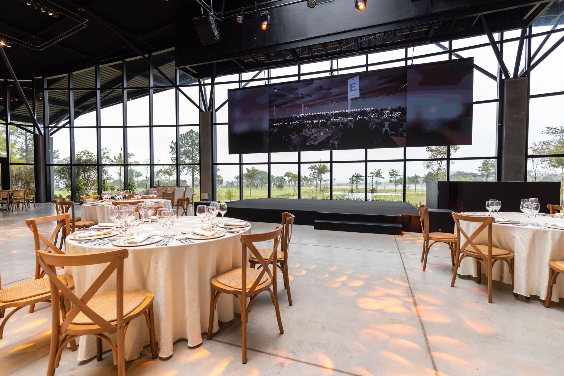 Elegante salón de banquetes con mesas redondas, mantelería blanca, sillas de madera y un gran escenario con pantalla.