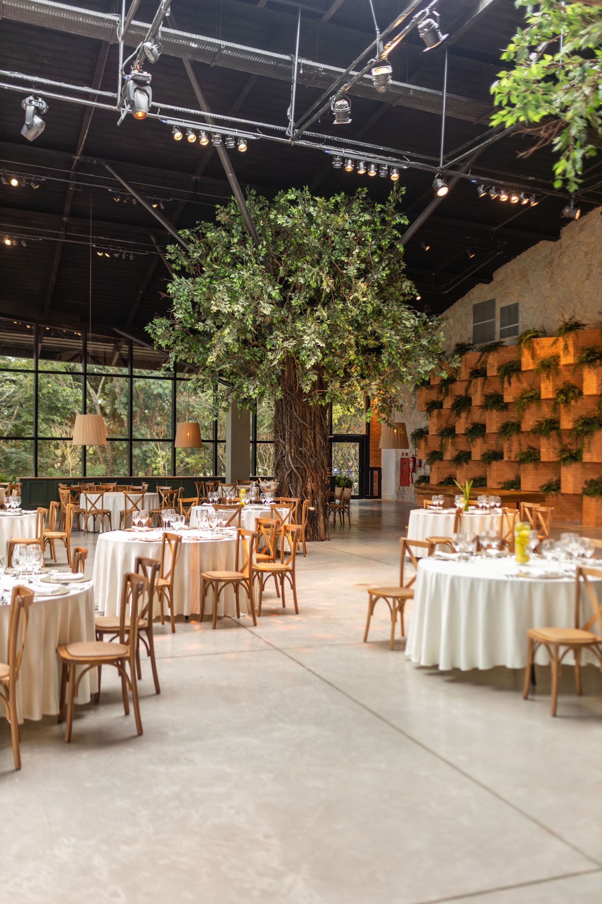 Elegante salón de eventos con mesas redondas, sillas de madera y plantas colgantes bajo un techo oscuro.