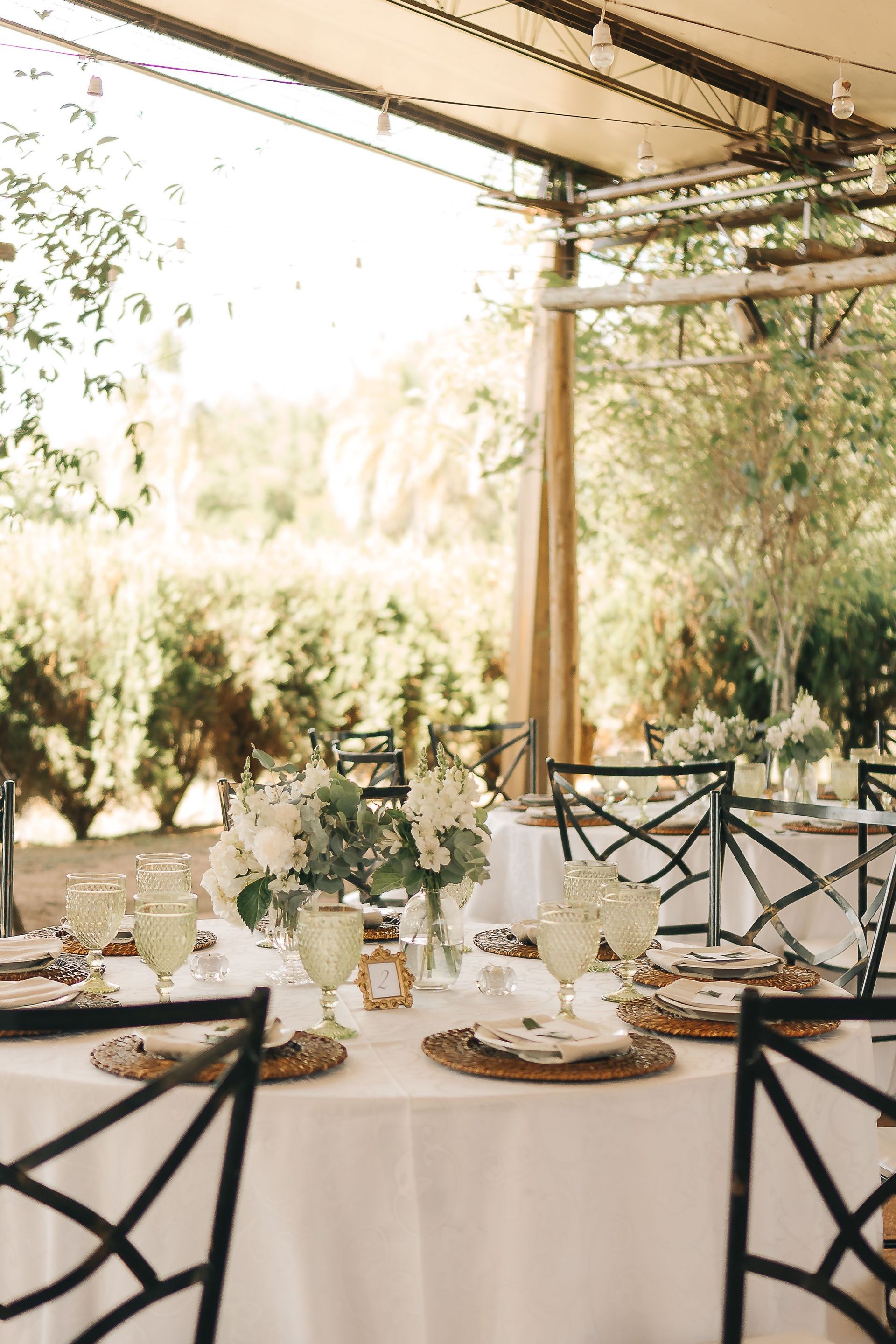 Elegante mesa de recepción al aire libre con manteles blancos, centros de mesa florales y sillas negras bajo un toldo.