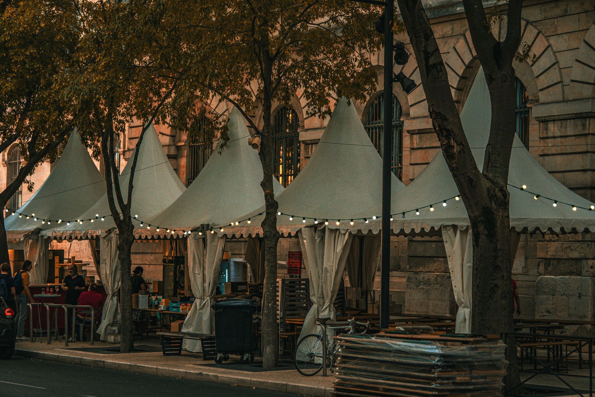 Puestos de mercado al aire libre con toldos blancos y guirnaldas de luces bajo los árboles al atardecer.