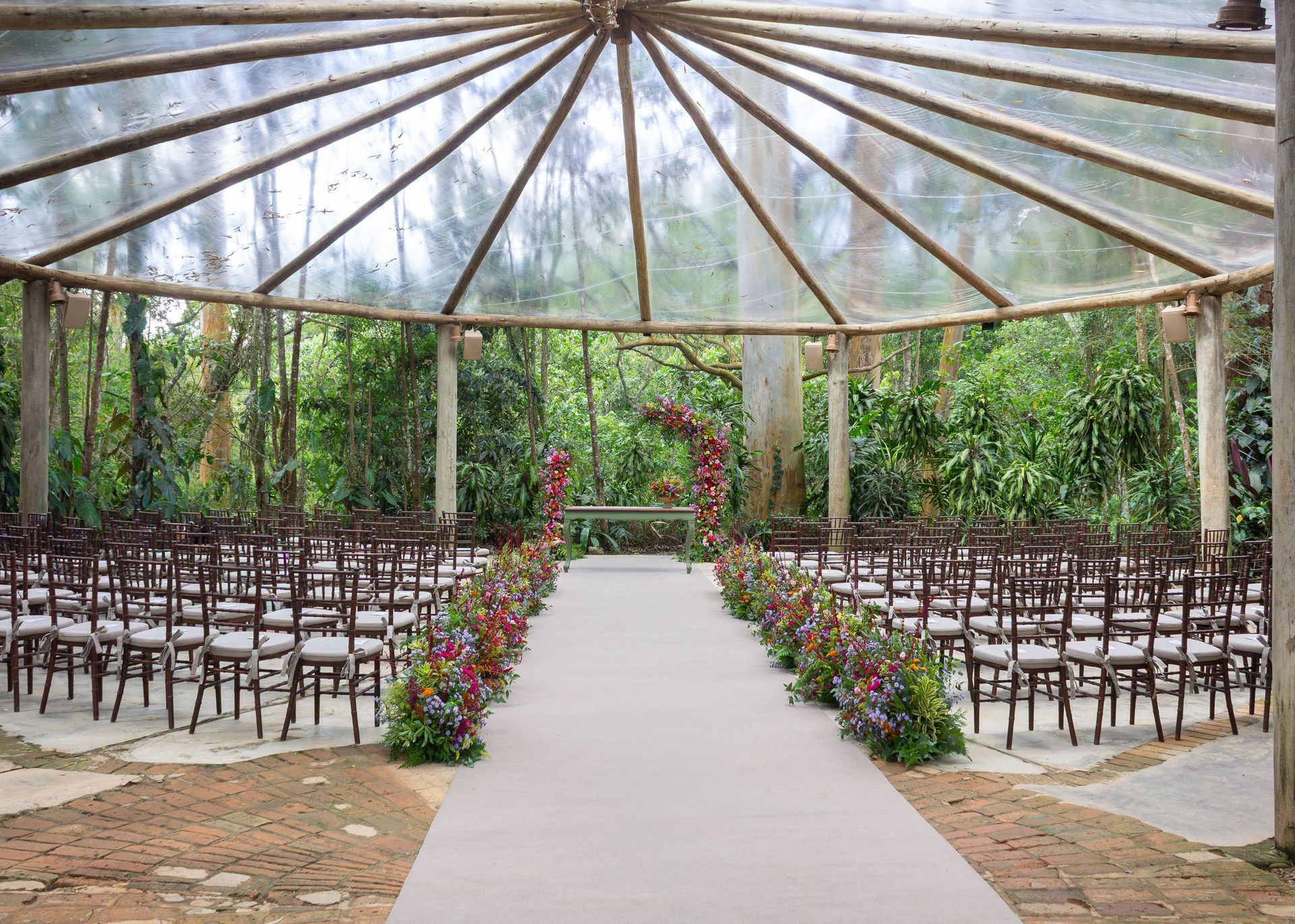 Montaje para ceremonia de boda al aire libre con filas de sillas bajo una pérgola rústica y un pasillo bordeado de flores.