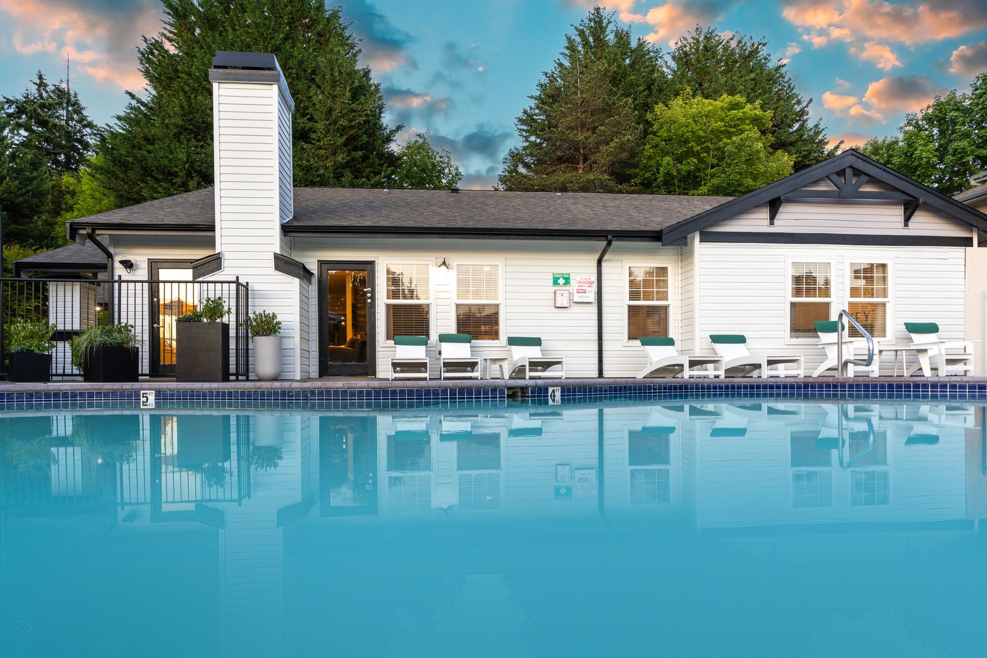 Poolside view of a white building with a dark roof, lounge chairs, and a swimming pool under a cloudy sky.