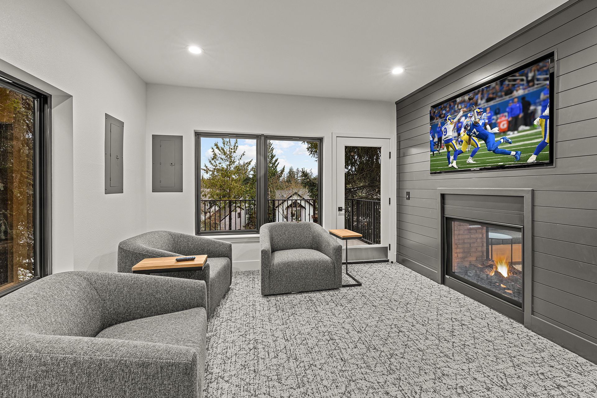Living room with grey armchairs, fireplace, and TV. Large windows offer a view of trees.