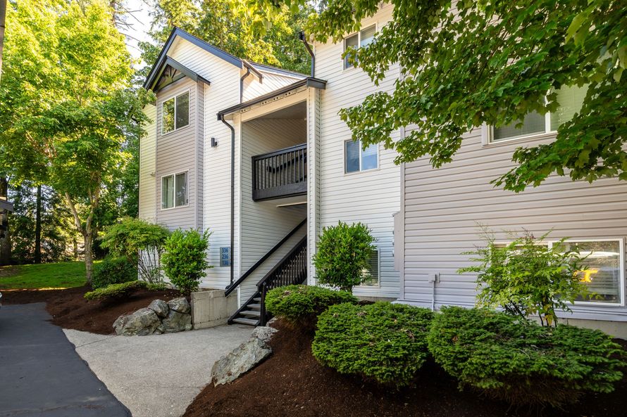 Apartment building entrance with white siding, stairs, and landscaped shrubs under trees