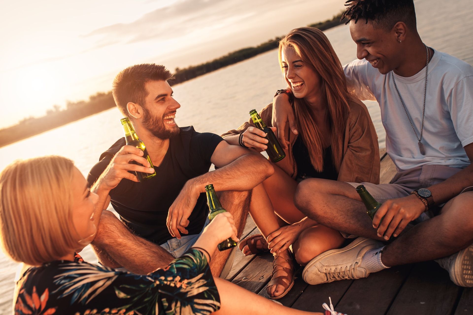 Friends relaxing on a dock at sunset, laughing and toasting with drinks by the water.