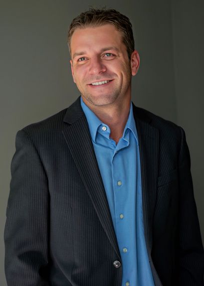 Smiling man in a dark blazer and blue shirt standing against a gray background