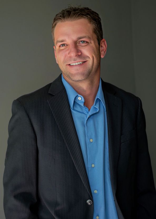 Smiling man in a dark blazer and blue shirt standing against a gray background