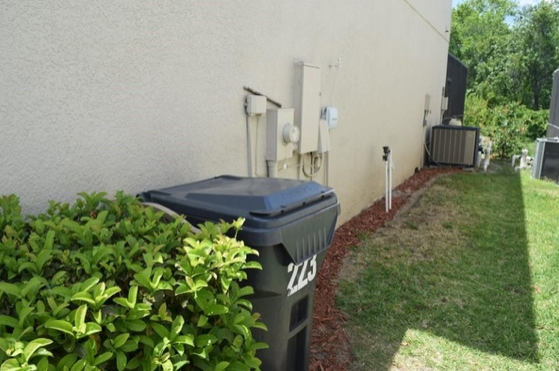 Side yard with trash bin, beige house wall, grass, bushes, and utility equipment.