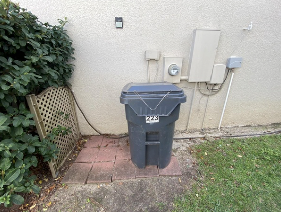 Black trash bin on a brick pad beside a house wall, with bushes and grass nearby.