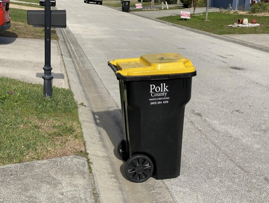 Black Polk County trash bin with yellow lid on a residential street curb