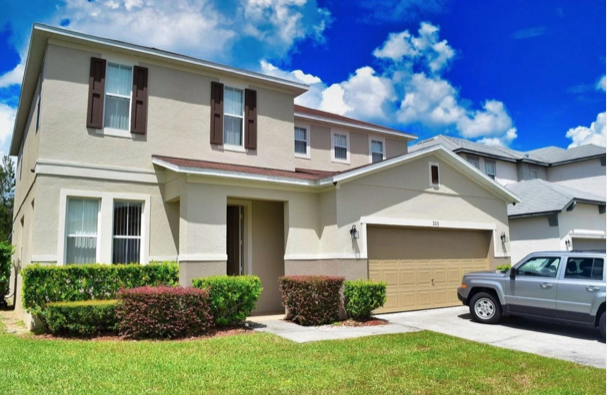 Two-story beige suburban house with white trim, garage, driveway, and a gray SUV under a bright blue sky.
