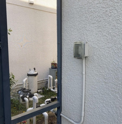 Outdoor utility equipment beside a stucco wall, seen through a metal fence.