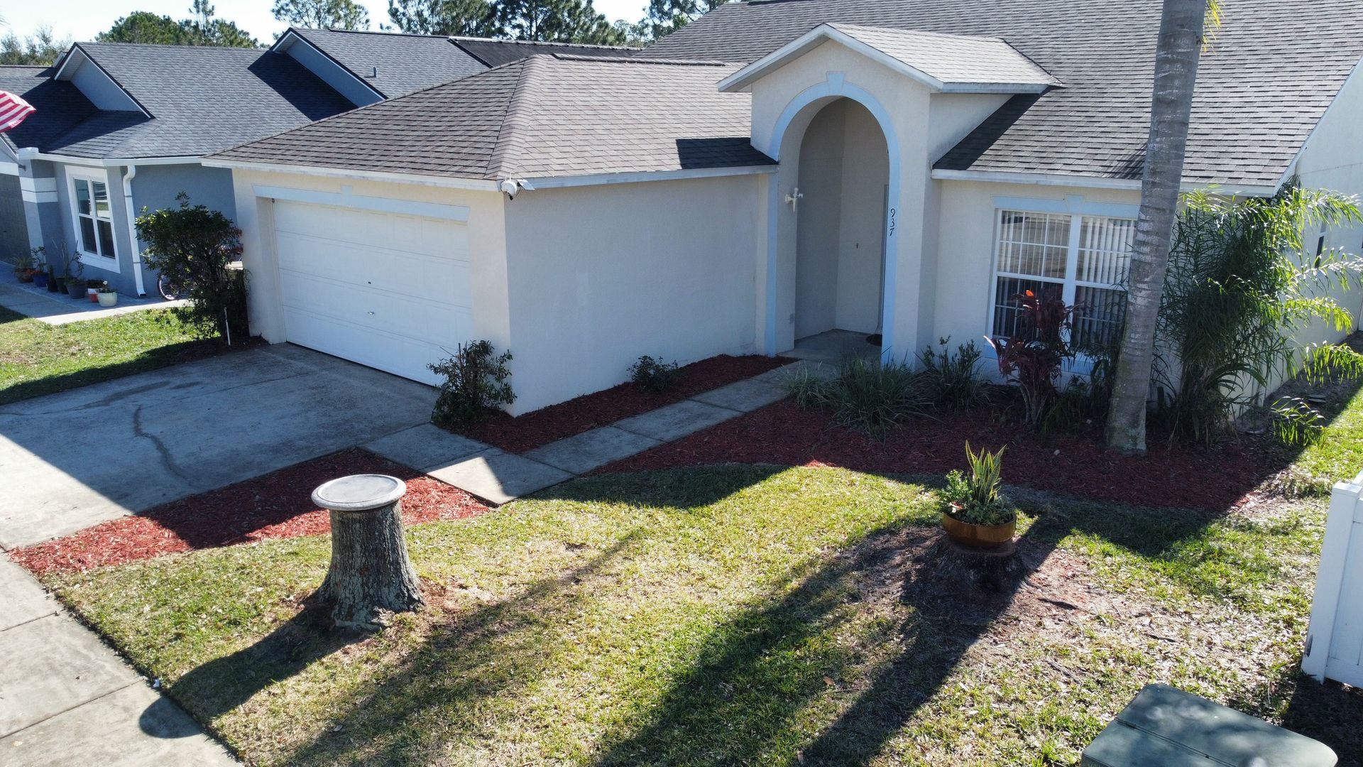 Suburban house with two-car garage, arched entryway, and a sunlit front yard with a tree stump.