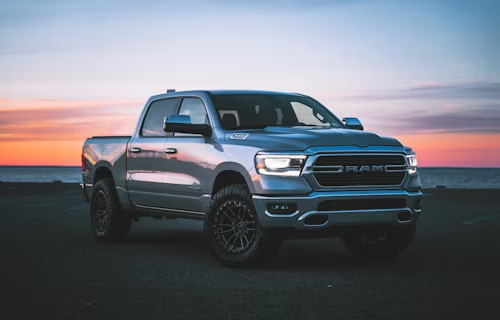 Silver Ram truck parked on asphalt with ocean and sunset in background.