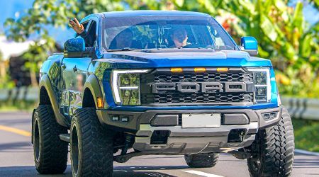 Blue Ford Raptor truck driving down a road, with a person waving from the window.