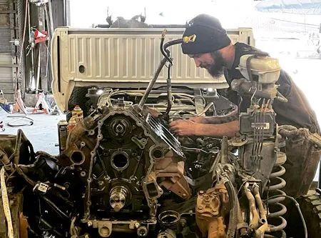 Mechanic working on a truck engine in a garage. Black beanie, focused expression, using tools.