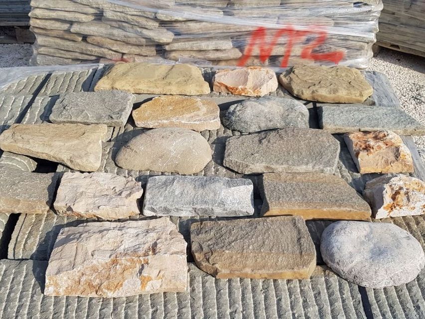 Campioni di rivestimento in pietra impilati nei toni del marrone chiaro, grigio e beige, con marcatura rossa, su un pallet all'aperto.