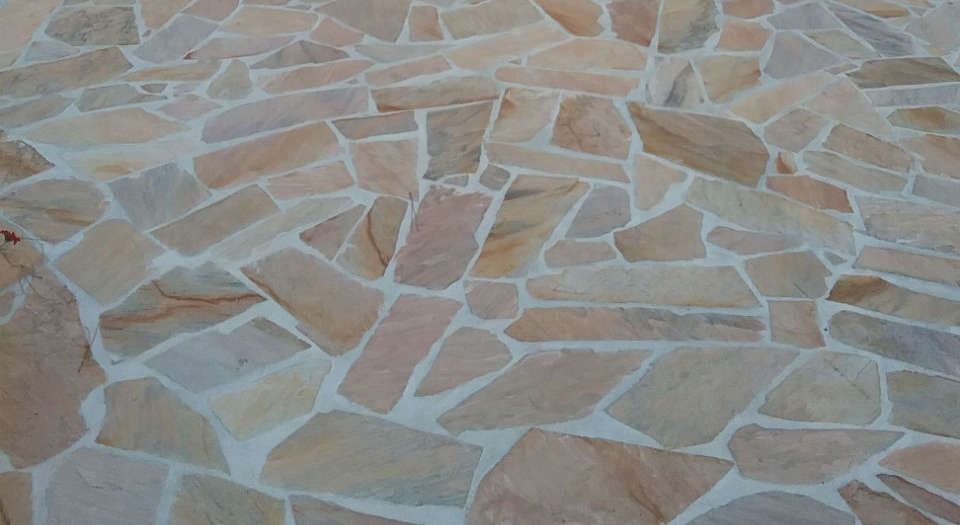 Primo piano di un pavimento in piastrelle di pietra color marrone chiaro e beige con tasselli di mosaico irregolari e fughe chiare.