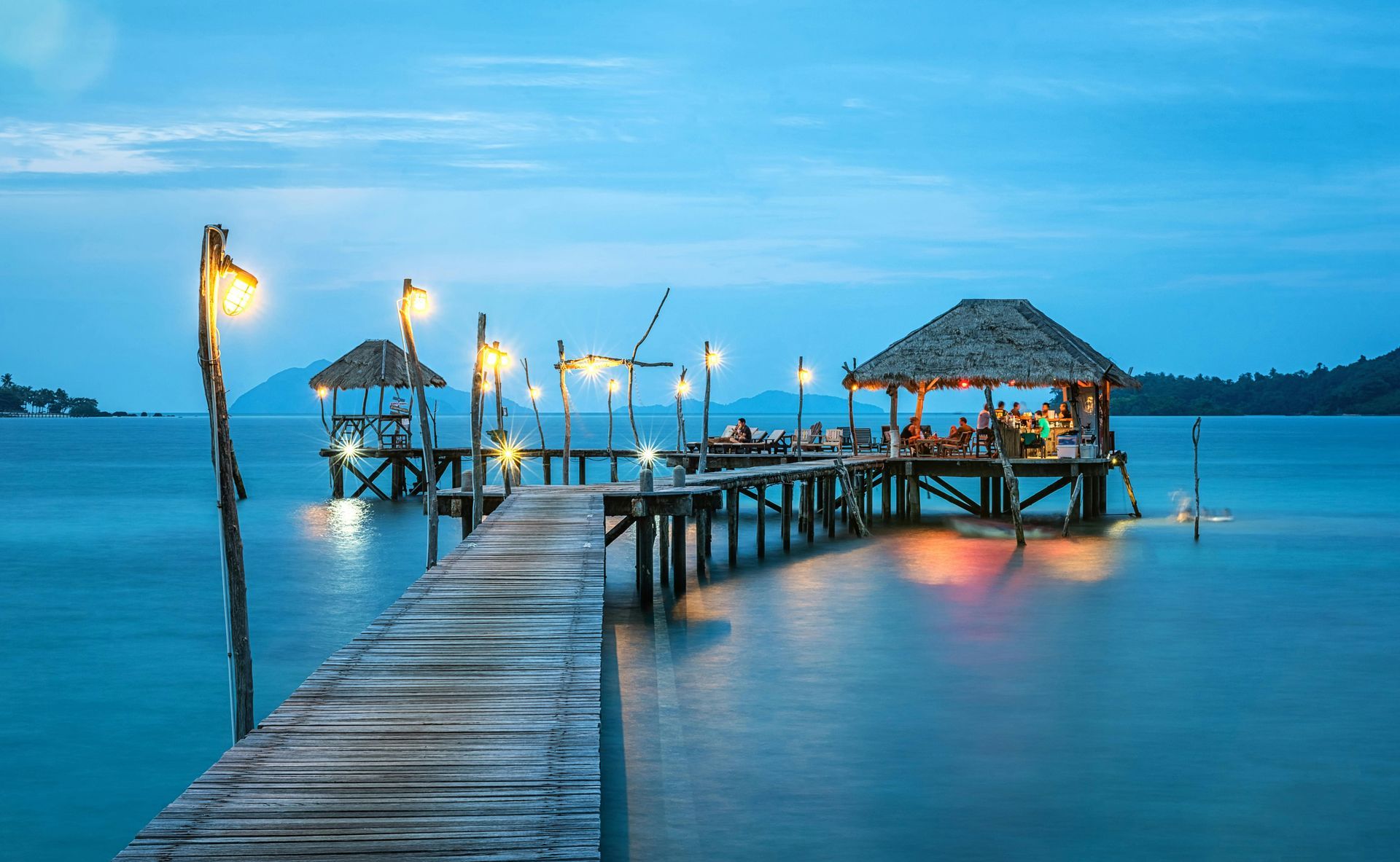 Wooden pier with thatched roof structures extending over calm blue water at dusk, lit by string lights.
