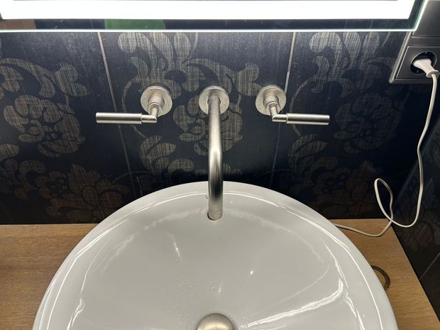 Bathroom sink with chrome faucet and handles against dark patterned wallpaper under a mirror light