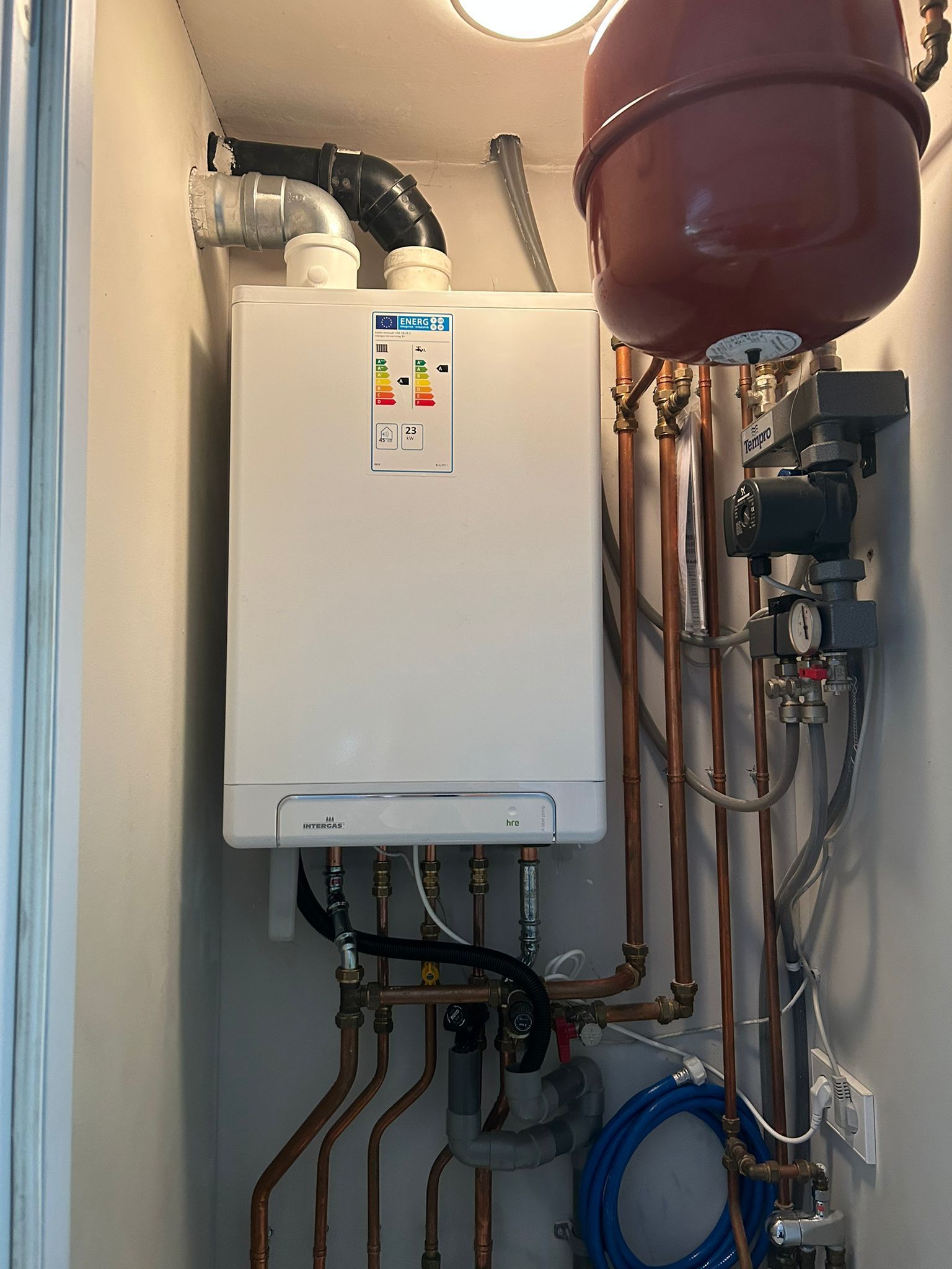White wall-mounted boiler in a utility room with exposed copper pipes and a blue hose reel.