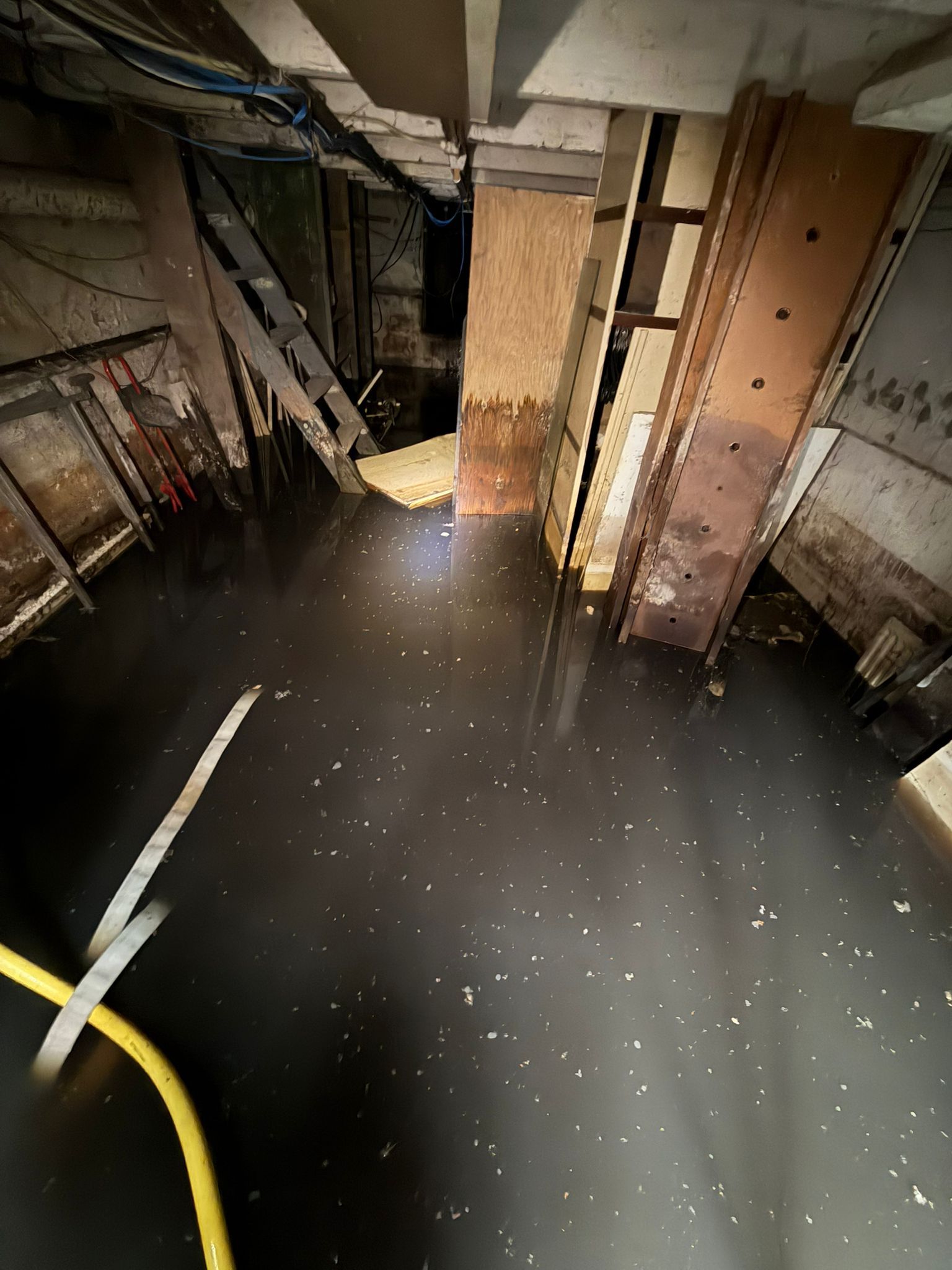 Flooded basement with dark water, exposed pipes, and a yellow hose in the foreground