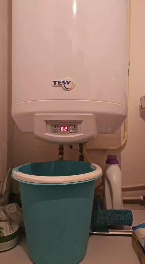 White TESY water heater over a blue bucket catching dripping water in a bathroom.