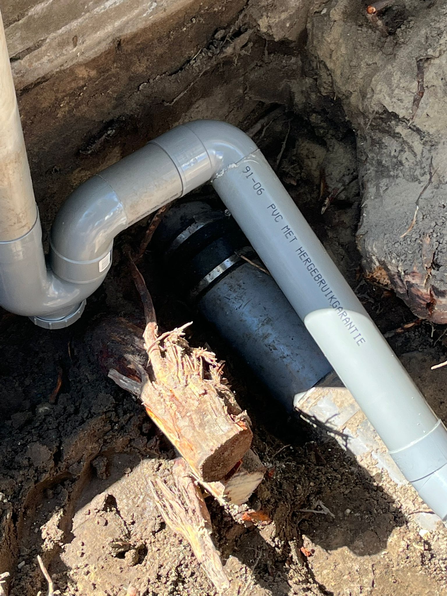 Underground plumbing pipes in a dirt trench, showing white PVC and black pipe connected near a foundation.