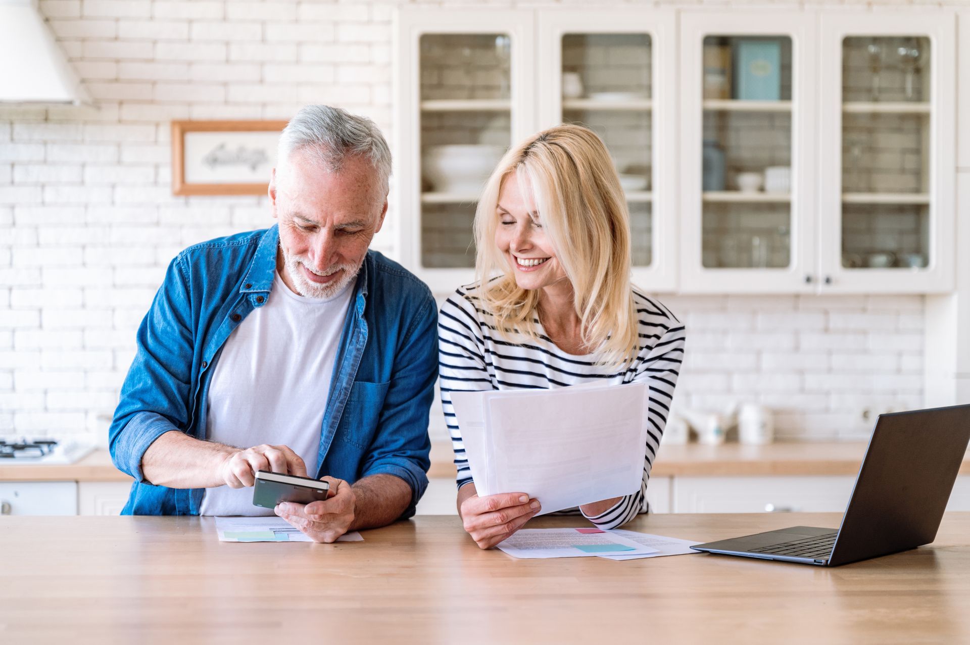 a man and a woman are sitting at a table in a kitchen looking at papers and a laptop