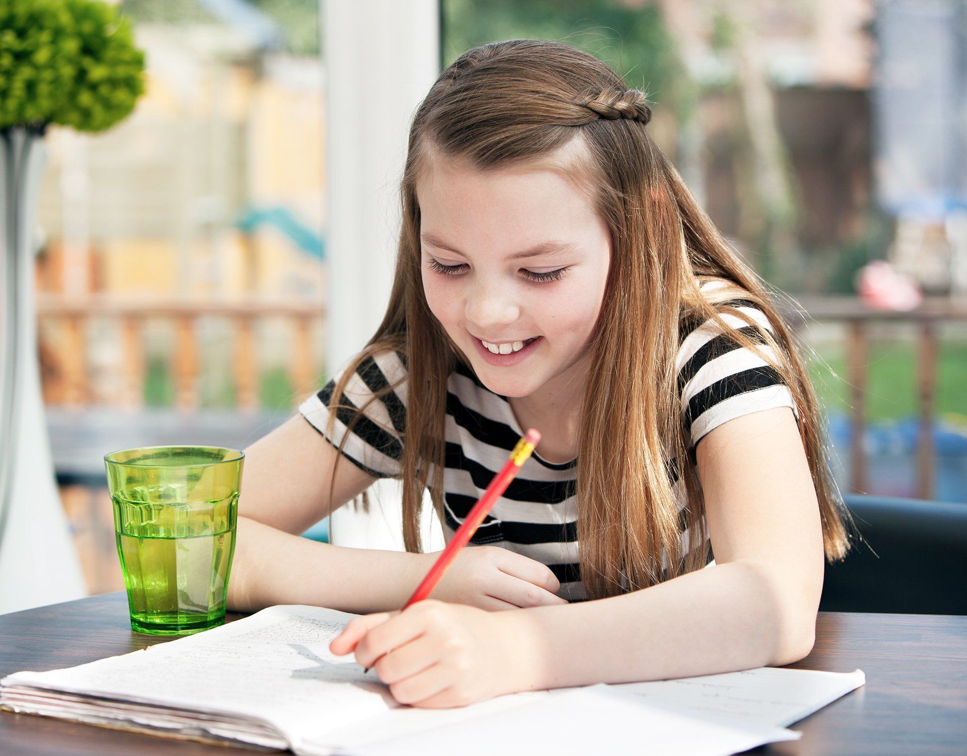 Girl with brown hair, smiles, writing in notebook at a table, glass of green liquid, striped shirt, pencil.