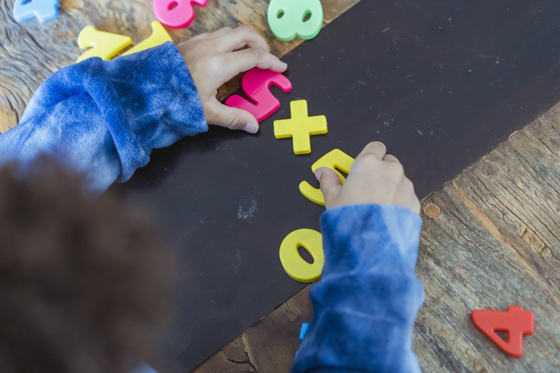 Child's hands arranging colorful foam numbers and a plus sign on a dark surface.