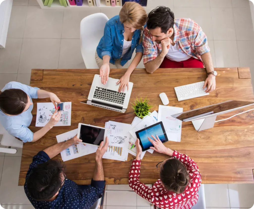 A group of people are sitting around a wooden table with laptops and tablets.