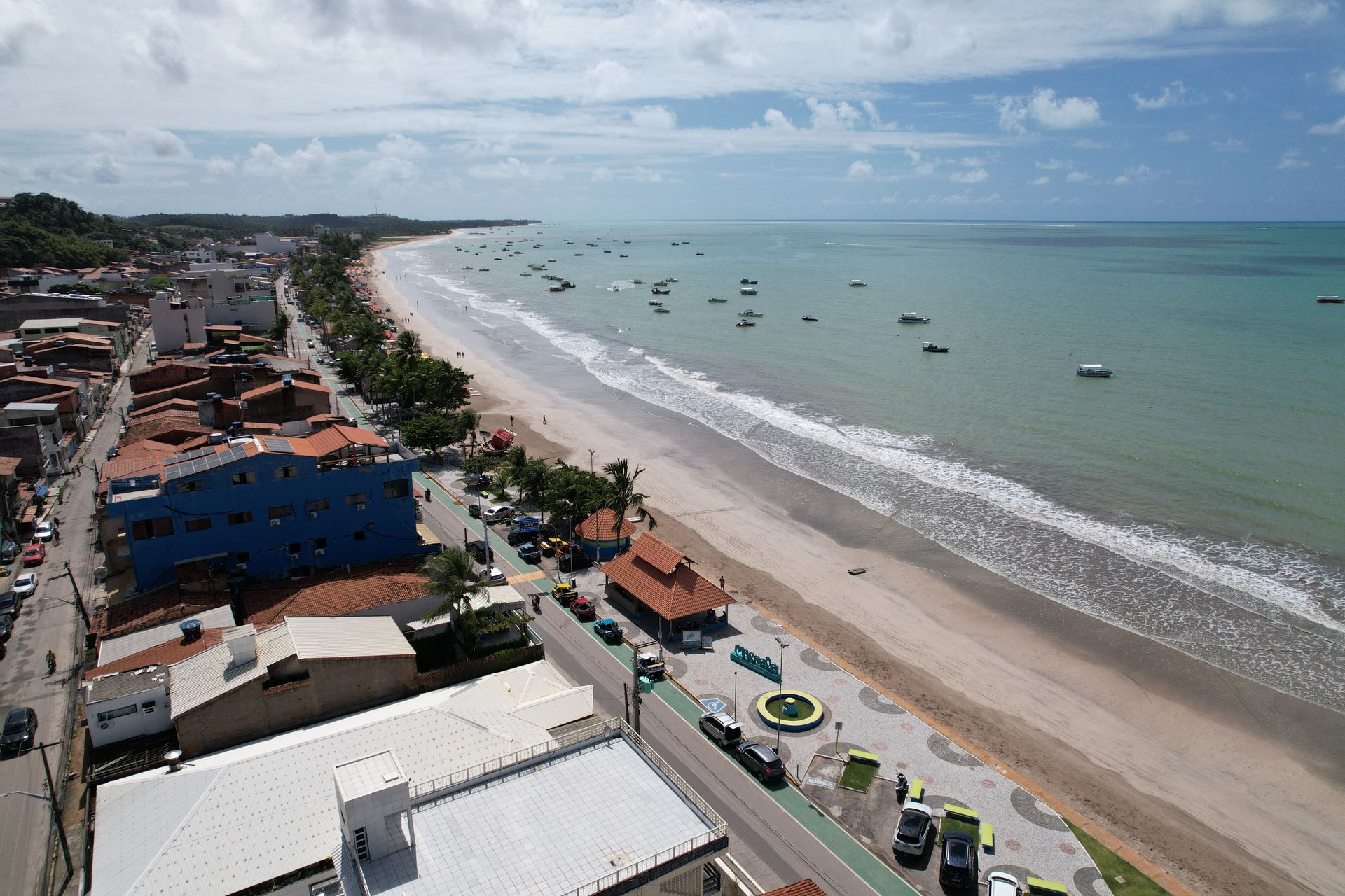 Vista aérea de uma cidade costeira com uma longa praia de areia, oceano azul e barcos ao largo.